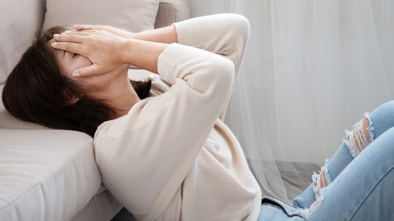 A woman seemingly in distress sitting on the floor with her hands over her face and her head laid back on a coach