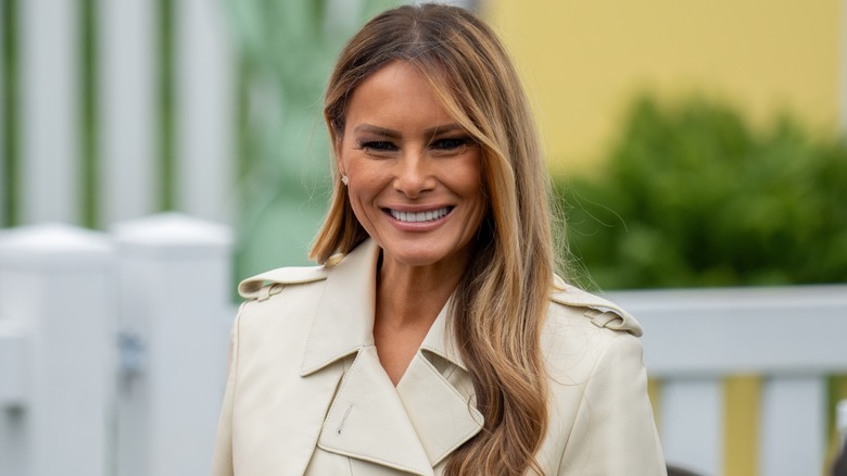 Melania Trump smiling in 2025 with a cream trench and her classic glossy waves