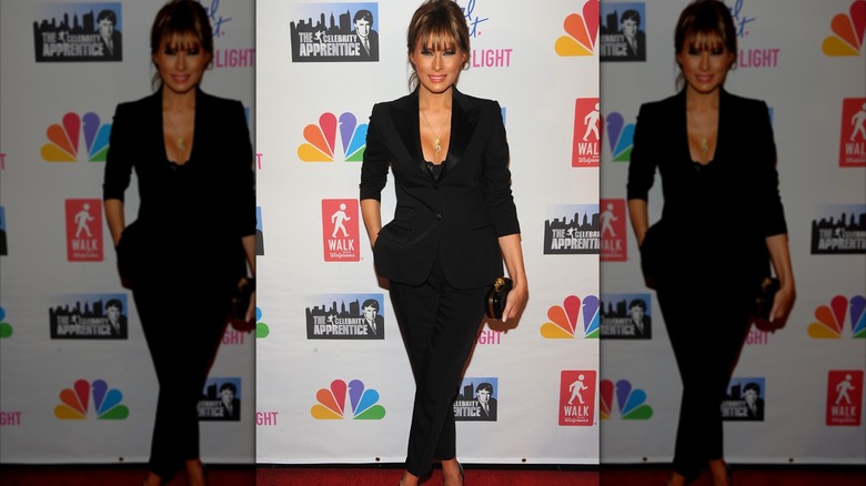 Melania Trump in a dipping black pantsuit standing with her legs crossed while posing for a photo