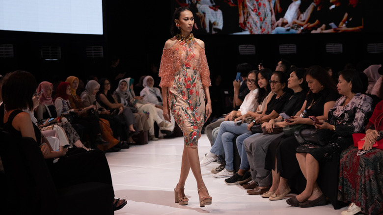 A model walking a runway in a floral dress with a sheer pink top