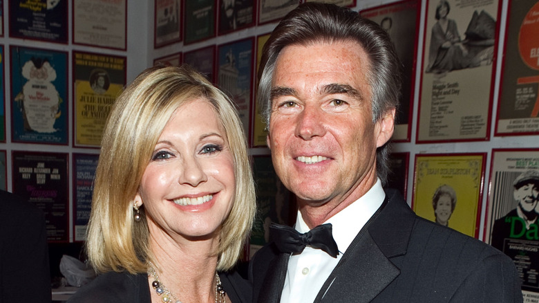 Olivia Newton-John smiles while posing next to second husband, John Easterling in front of a poster gallery wall.