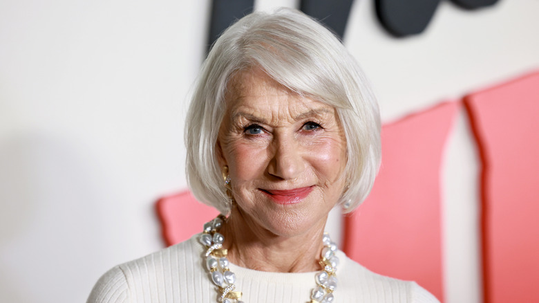 Helen Mirren posing at a red carpet event wearing a white dress and large necklace.