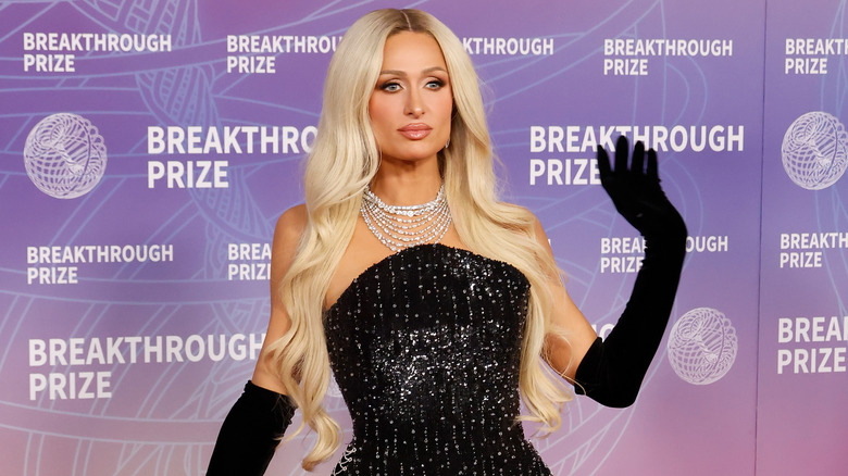 Paris Hilton waving at a red carpet event wearing black gloves, a black dress, and sparkly statement necklace.
