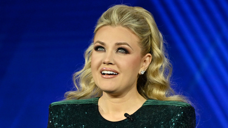 Erika Kirk with dramatic makeup and pageant-style hair, speaking into a microphone