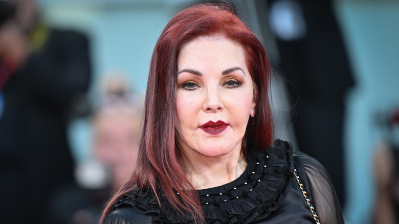 Priscilla Presley sports red hair and a full glam makeup look, including dark burgundy lipstick.