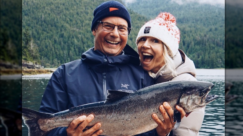 Sarah Chalke sports a wide smile in front of a water landscape next to her boyfriend Michael Leckie holding a fish