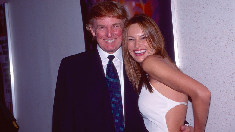 Donald Trump in a black suit and Melania Trump in a white dress, smiling for a photo at a movie premiere