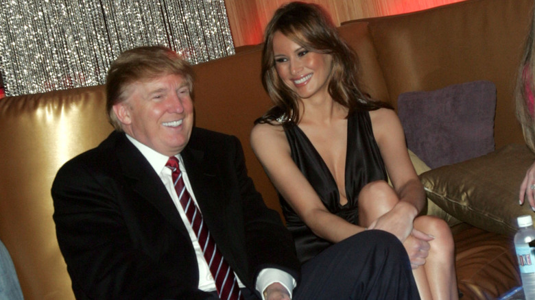Melania Trump wearing a black dress with crossed legs smiling at Donald Trump who is wearing a black suit