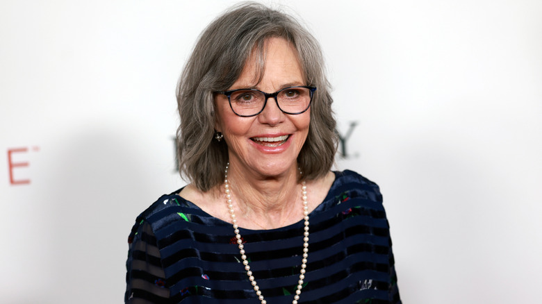 Sally Field smiling at a red carpet event wearing a sparkly black top and long pearl necklace.