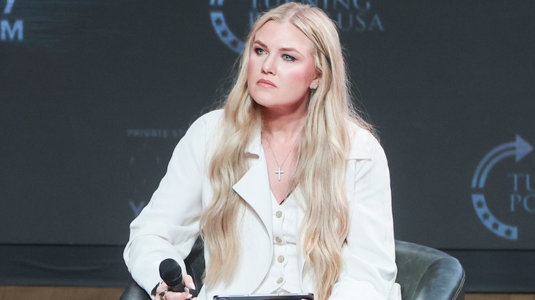 Erika Kirk is seated at a Turning Point USA event. This image shows off her ultra-long blonde extensions. She is a woman with fair skin, and is wearing a white suit jacket and matching vest.