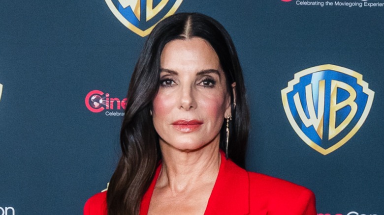 Close-up image of Sandra Bullock. She is an older woman with fair skin and long dark brown hair. She is wearing a bright red suit jacket.
