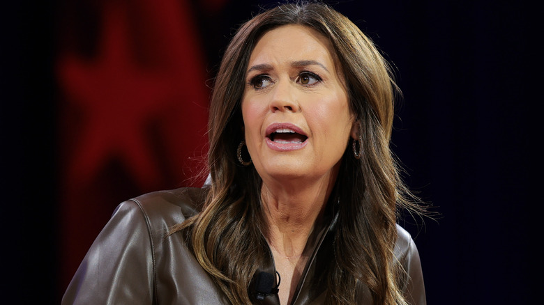 Sarah Huckabee Sanders speaking with long, flowing brunette hair and a brown jacket