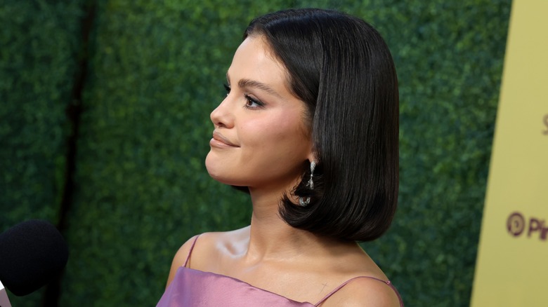 Selena Gomez poses in front of a green backdrop at an event, with a sleek bob hairstyle.