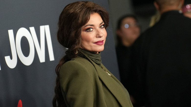 Shania Twain wears an olive green turtleneck and matching blazer with deep brunette hair styled in a braid.