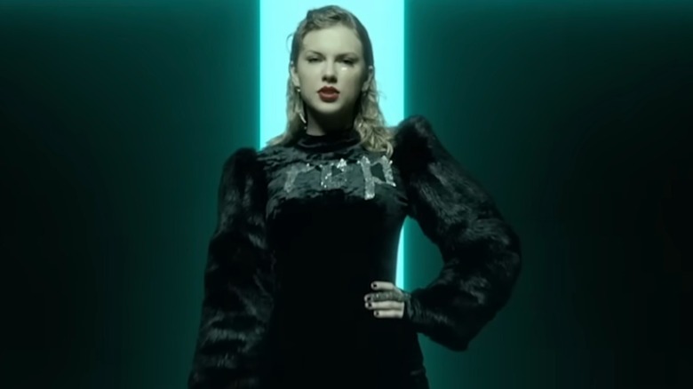 Taylor Swift wears a black fur bodysuit with long sleeves and the word "REP" embellished across her chest in silver. Her hair is tucked behind her ears, and she is wearing red lipstick.