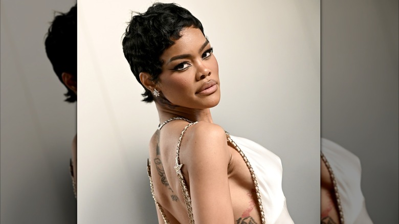Teyana Taylor posing at a 2026 Oscars event. She is looking over her shoulder and wearing a backless cream silk gown with gold chain accents.