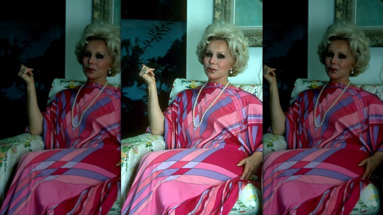 Eva Gabor poses wearing a pink, purple, and blue dress on a couch with her hand up.
