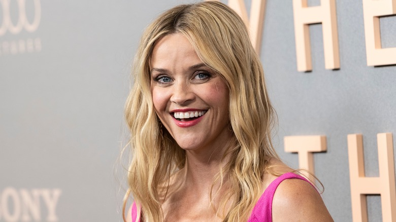 Reese Witherspoon smiling on the red carpet with red lipstick and a pink dress
