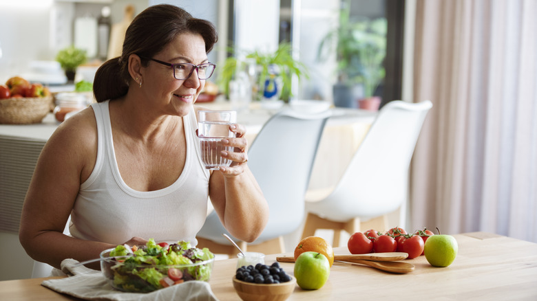 A middle-aged woman drinks water as she sits at a table of fruits and vegetables