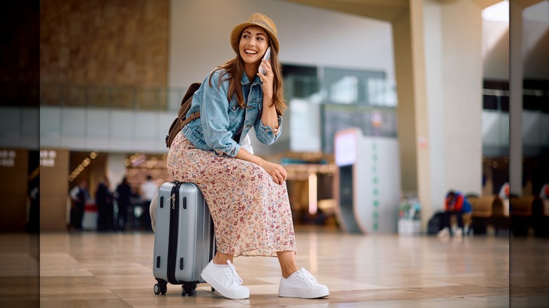 A person sitting on a suitcase on the phone wearing a dress, denim jacket, and white sneakers