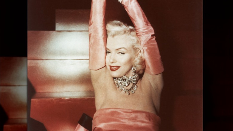 Marilyn Monroe in a jeweled dress with fur jacket leaning against a dresser