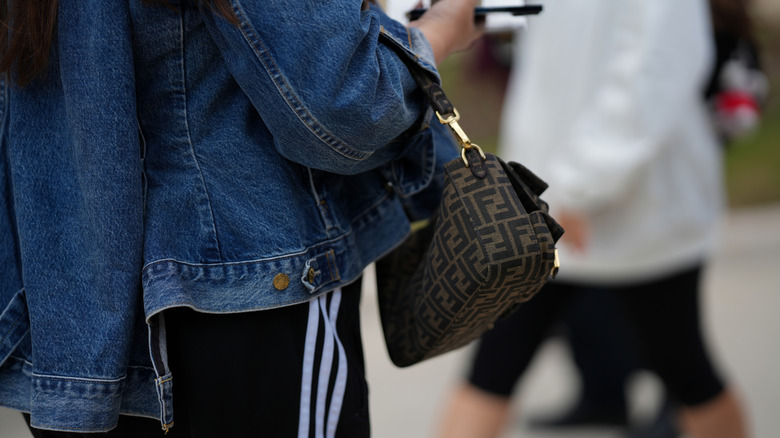 A person holding wearing track pants, a jean jacket and accesorizing with a Fendi bag