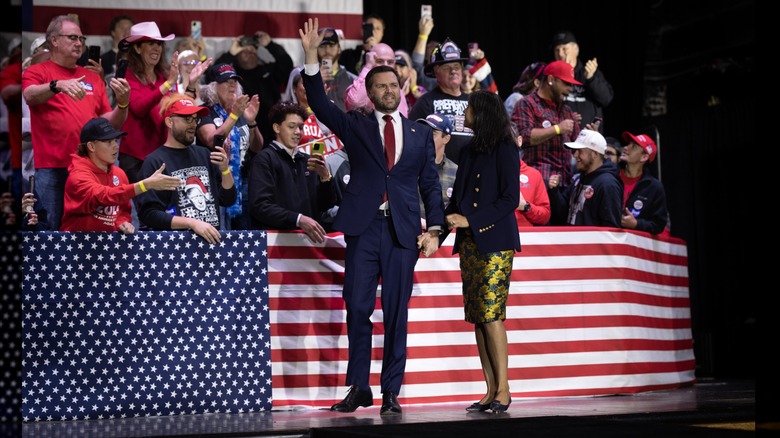 Usha Vance stands beside JD Vance on the campaign trail. She is wearing a dark blue blazer, a blue and yellow floral midi skirt, and black kitten heels
