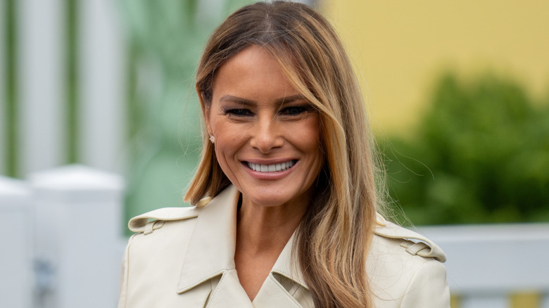 Melania Trump smiling in a tan trenchcoat