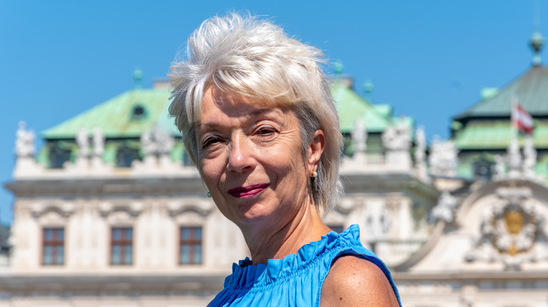 An older woman in a blue outfit and grey textured feathered hair poses for a picture