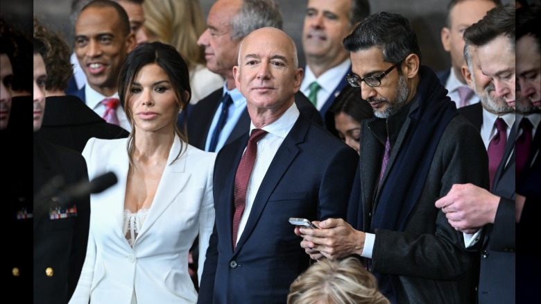Full image of Lauren Sanchez Bezos and Jeff Bezos at the 2025 Presidential inauguration. She is wearing a white blazer with a white lacy bra top underneath. He is wearing a dark navy suit with a white shirt and dark red tie.