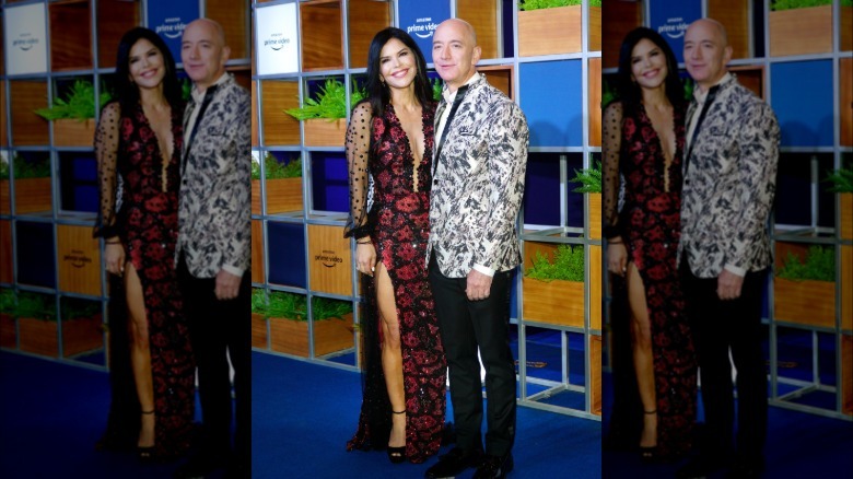 Full image of Lauren Sánchez Bezos and Jeff Bezos. Lauren is wearing a long red and black beaded gown with a high split and low neckline. Jeff is wearing a patterned suit jacket and black pants.